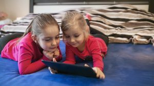 Two girls sitting on a couch enjoying screen time, watching cartoons on a tablet.